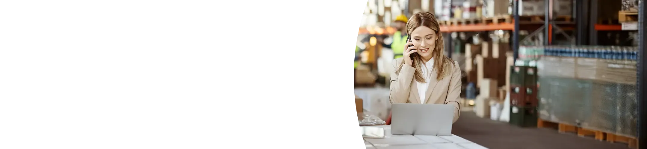 A smiling female manager in a business suit is talking on a phone and working on a laptop on top of wrapped pallets in a busy warehouse.